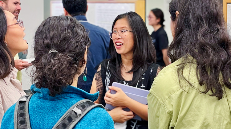 A group of students and researchers engage in lively conversation during an academic poster session. One person at the center smiles while holding a notebook, surrounded by others wearing casual attire and backpacks, with research posters visible in the background.